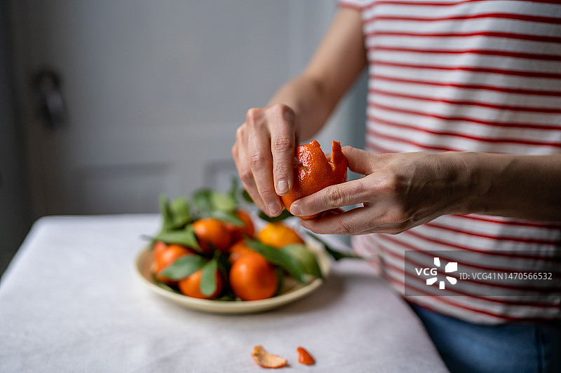 穿着条纹T恤的特写女人，在桌子上的盘子里剥着带有绿色新鲜叶子的橘子图片素材