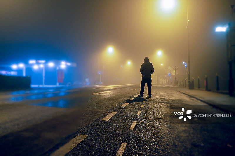 一个戴着兜帽的神秘人物，背对着镜头，站在城市一条空旷的道路上。一个怪异的冬夜图片素材