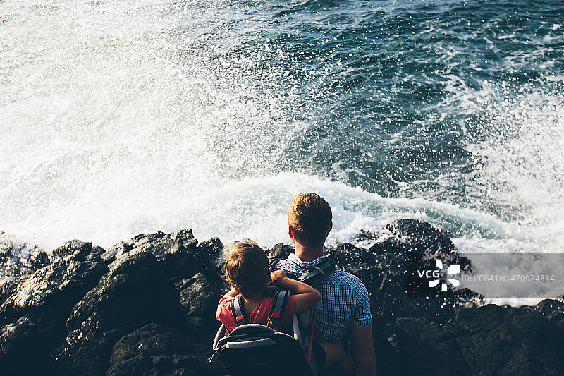千禧一代男人带着坐在婴儿背带里的男孩在火山海滩上徒步旅行，海景图片素材