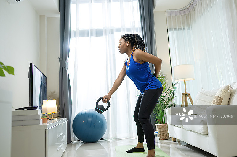 壶铃摇摆锻炼燃烧卡路里并增强力量。非洲裔美国妇女在家中锻炼期间，通过智能电视观看在线课程，进行壶铃举重和深蹲。图片素材