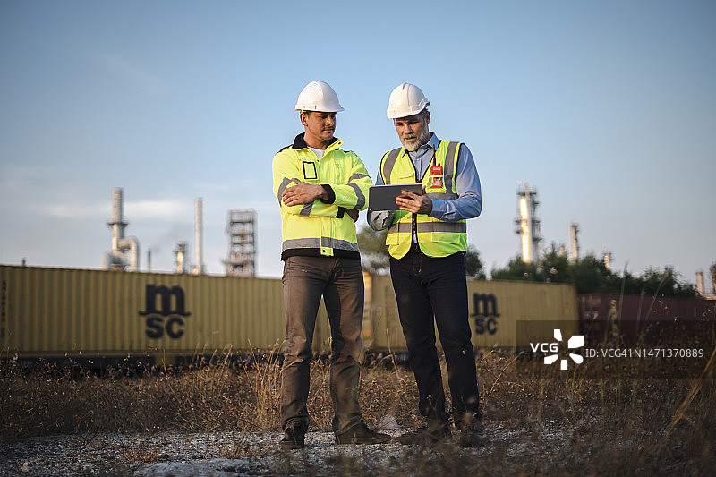 穿着反光工作服的白人工程师和初级白人工程师。站在炼油厂前，咨询输油管道的铺设图片素材
