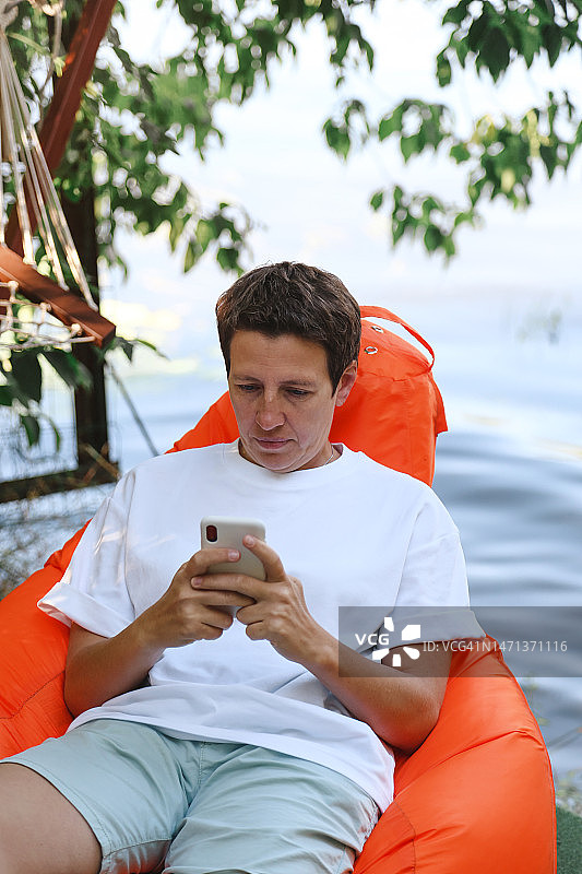 中年女商人使用手机，在模糊的湖岸背景下，坐在橙色豆袋椅上休息图片素材