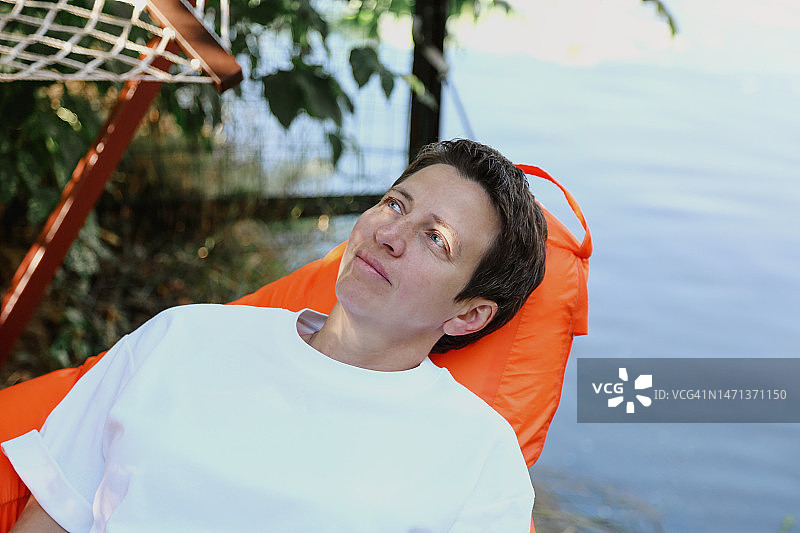 中年女商人在橙色豆袋椅上放松，背景是模糊的湖岸和吊床网图片素材