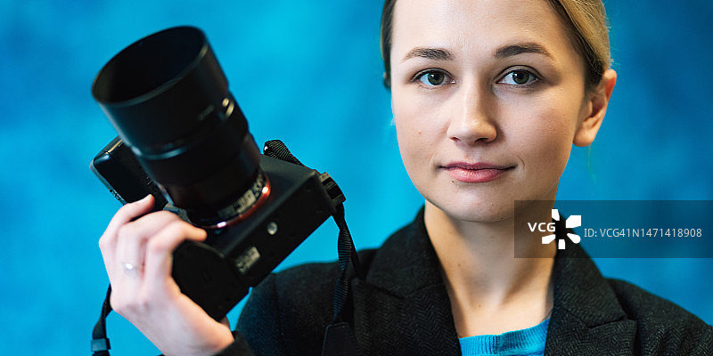 摄影师在蓝色背景工作室使用微单数码相机拍摄的女性肖像。该女人身穿黑色商务夹克和蓝色T恤，室内生活方式概念摄影。图片素材