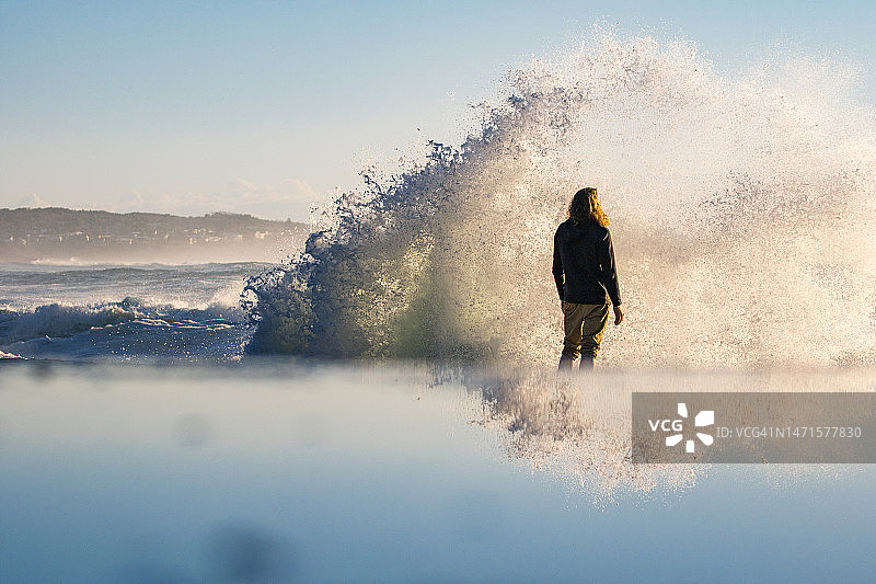 男人站在水边沉思，海浪拍打着岩石，沐浴在温暖的金色晨光中图片素材