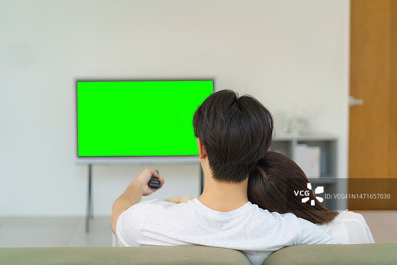 亚洲夫妇在客厅的沙发上看电视，女人靠在男人的肩膀上，象征着承诺和爱图片素材