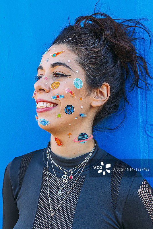 面部贴着未来主义服装、星系和宇宙贴纸的西班牙裔千禧一代微笑女性，蓝色墙壁背景图片素材