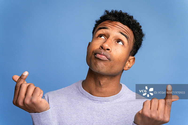 仰视休闲装扮的年轻非裔美国男子，做出数钱手势，看向蓝色背景前的镜头图片素材
