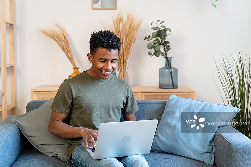穿着休闲装的乐观的非洲裔美国男性自由职业者坐在沙发上，浏览笔记本电脑，在家中远程工作图片素材