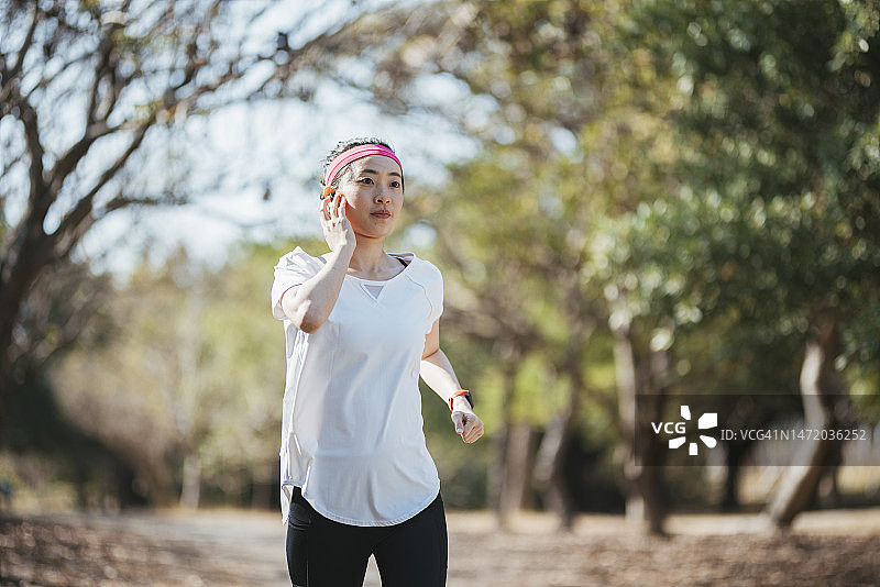 一位亚洲女性在户外慢跑，同时使用蓝牙耳机听音乐或参与远程指导课程。图片素材