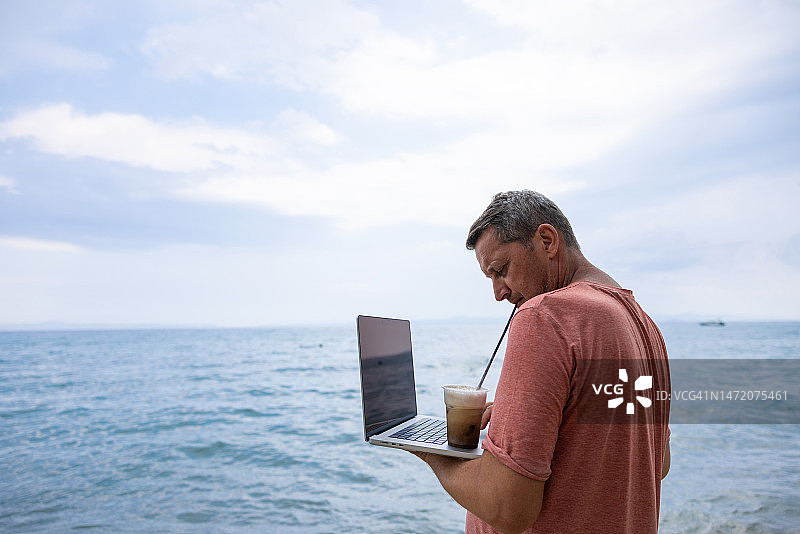 白人男子在海滩上使用笔记本电脑，同时喝着冰咖啡图片素材