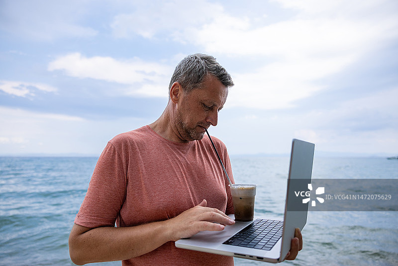 白人男子在海滩上使用笔记本电脑，同时喝着冰咖啡图片素材