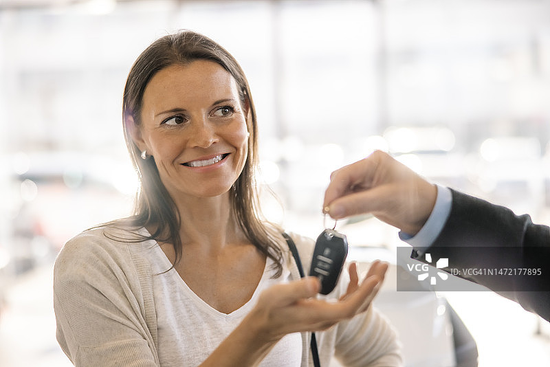 特写：女人从汽车销售员手中接过新车钥匙，笑容满面，两人站在汽车经销商店内图片素材