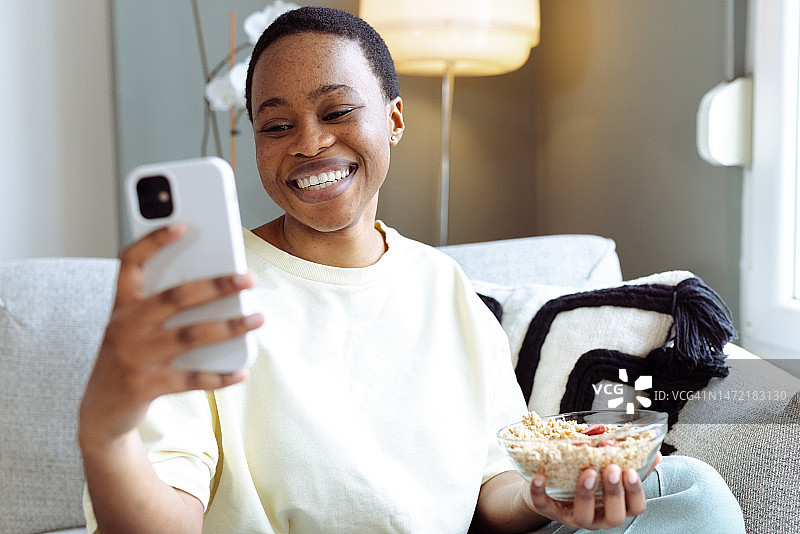 快乐的非裔美国年轻女性吃早餐并用智能手机上网图片素材