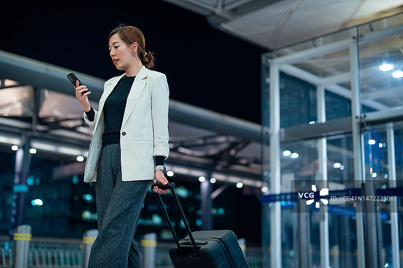 亚洲年轻女商人拉着行李箱，看着智能手机从机场走出来，走在航站楼里。商务旅行。旅游和度假概念。生活方式和技术。旅途中的商务图片素材