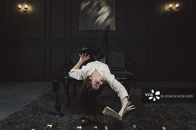 一位带有哥特式妆容和红褐色头发的神秘女孩，穿着灰白色衬衫，在黑暗的哥特式房间里摆姿势，手里拿着镀金餐具和书图片素材