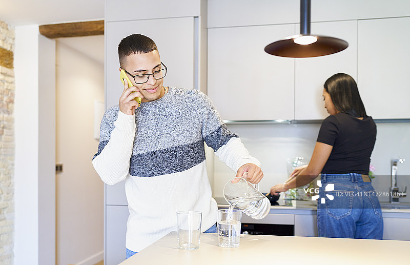戴眼镜的男人在打电话，倒水，背景是一位正在做饭的短发女人图片素材