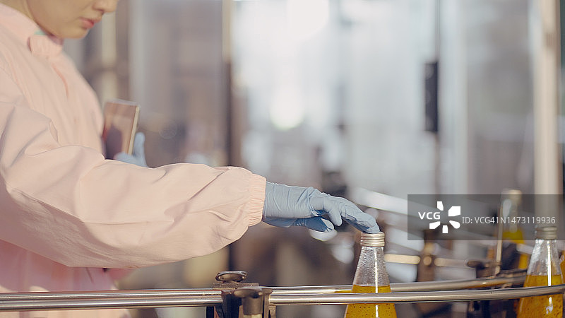 女食品科学家或营养师或质检员，戴发网和无菌服，在工厂生产线上检查灌装到瓶中的果汁质量。员工在制造厂瓶装果汁图片素材