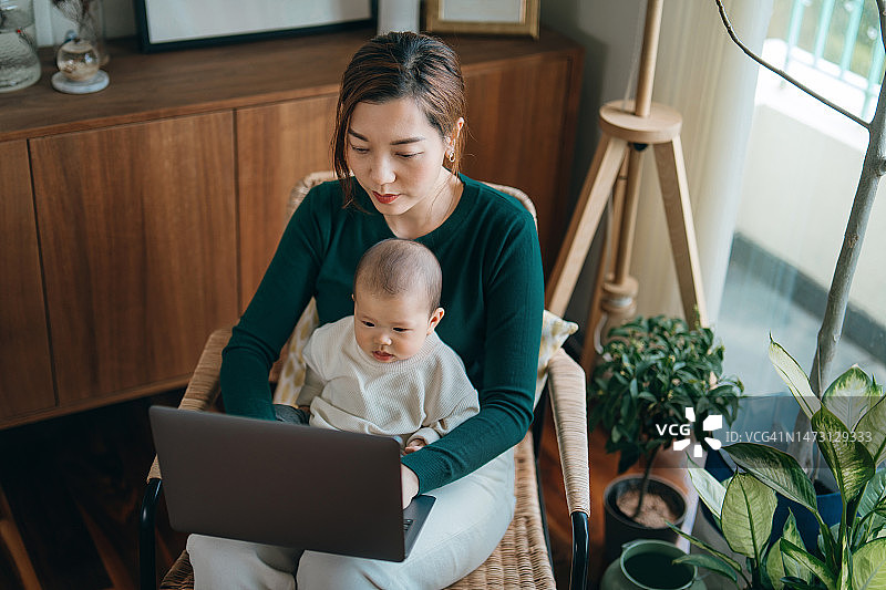 年轻的亚洲母亲在家使用笔记本电脑工作，同时照顾坐在腿上的女儿，身兼数职的职业母亲兼顾工作和育儿图片素材