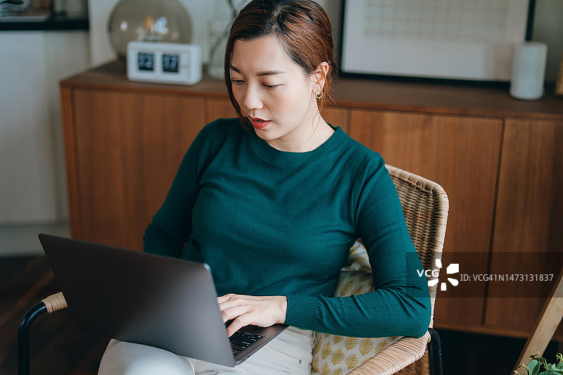 年轻的亚洲女人坐在舒适的家中的扶手椅上，使用笔记本电脑。自由职业者，自雇。在家工作。生活方式和科技图片素材