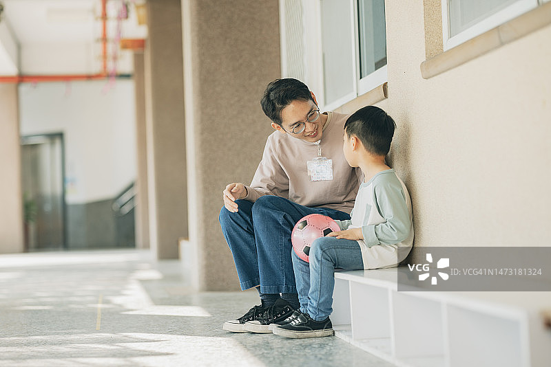 亚洲幼儿园老师坐在教室外面，与一个情绪低落的孩子交谈。她专心倾听并安慰孩子。图片素材