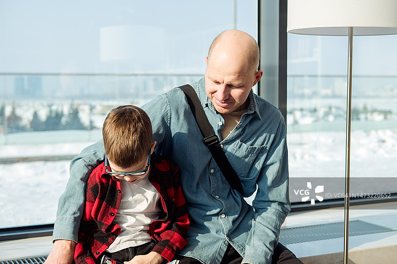 在图书馆大窗户旁的窗台上，一位39岁的秃头男人穿着牛仔衬衫和他的儿子一起读书图片素材