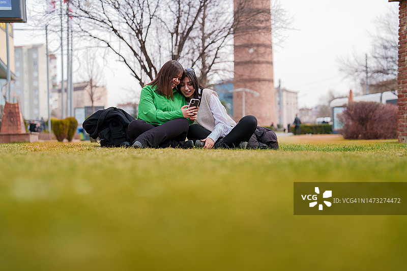 两位年轻女子坐在草地上使用移动设备图片素材