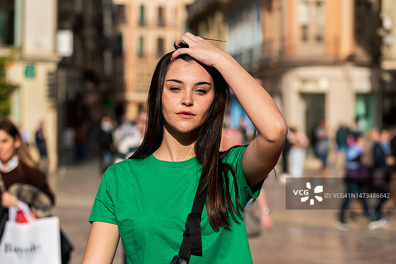 长发年轻女孩身穿绿色T恤，肩挎包走在城市街道上，抚摸着头发图片素材