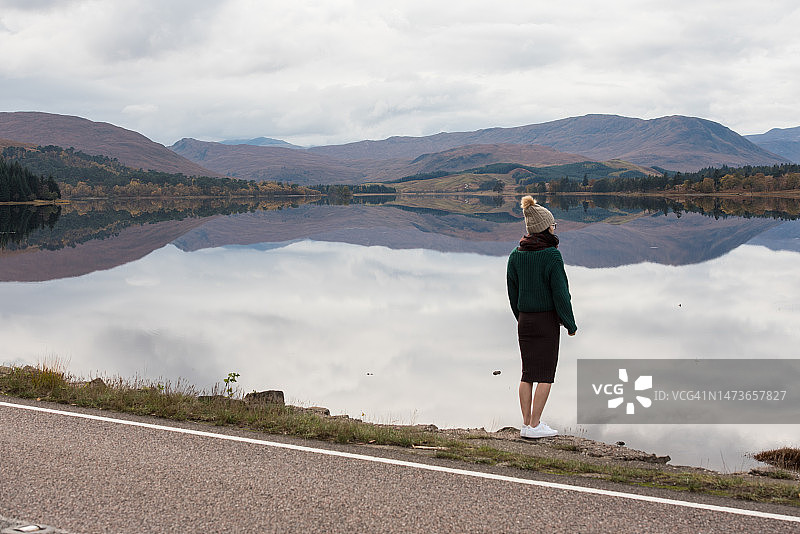 在英国苏格兰高地，一位戴着羊毛帽和围巾的女士站在一条可以俯瞰尼斯湖平静水面的道路旁图片素材
