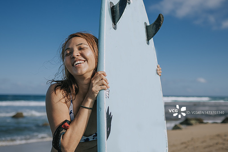 在海滩上，闭着眼睛微笑的亚洲女冲浪者站在冲浪板旁图片素材