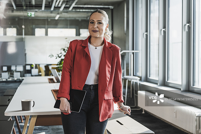 在办公室办公桌旁穿着红色西装的微笑女商人图片素材