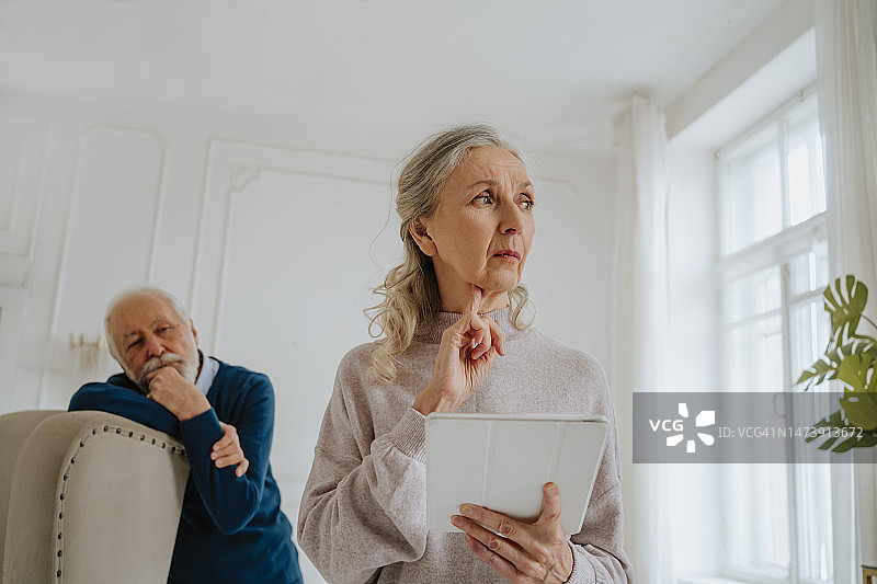 担忧的老年妇女在家中手持平板电脑，背景是她的丈夫图片素材