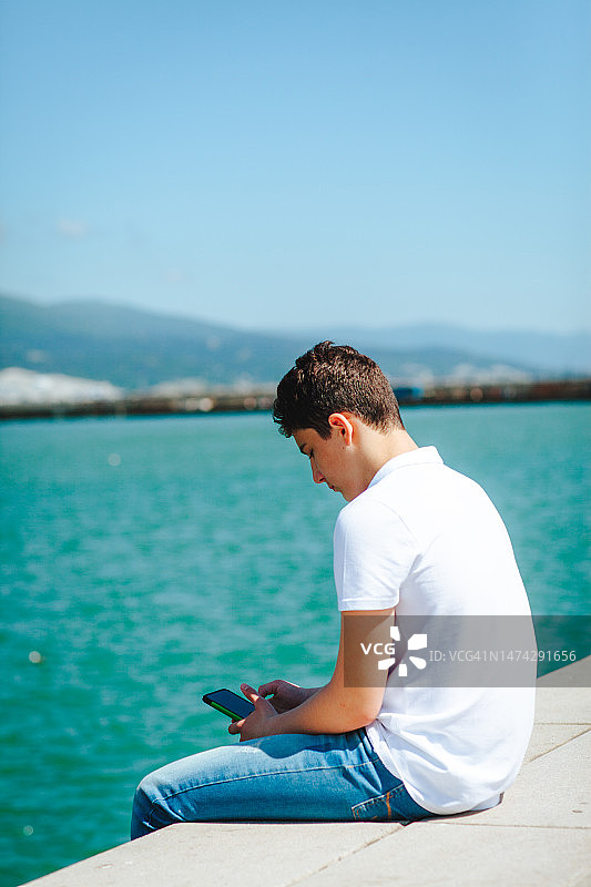 穿白色T恤男孩在海边玩手机图片素材