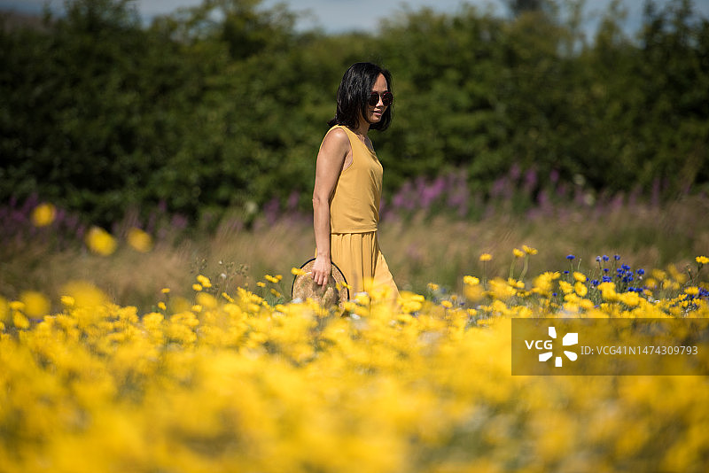一位年轻的亚洲女性戴着太阳镜，穿着黄色连体裤裙，走在苏格兰金罗斯的野生黄色花卉草地上，手里拿着草帽，沐浴在阳光明媚的夏日里图片素材