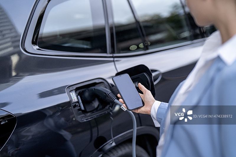 年轻女人在公共充电站为电动汽车充电并用手机支付图片素材