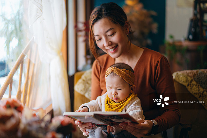 年轻的亚洲母亲在家给女儿读书，可爱的女儿坐在妈妈的腿上一起阅读故事书，促进儿童教育和发展。图片素材