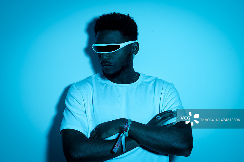 戴着未来主义时尚眼镜的年轻非裔美国男子，在蓝色霓虹灯下，双臂交叉。未来、现代技术、技术进步、独眼巨人眼镜的概念图片素材