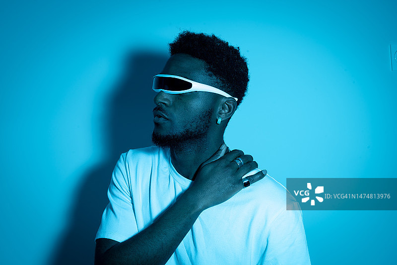 戴着未来主义时尚眼镜的年轻非裔美国男子，在蓝色霓虹灯下，双臂交叉。未来、现代技术、技术进步、独眼巨人眼镜的概念图片素材