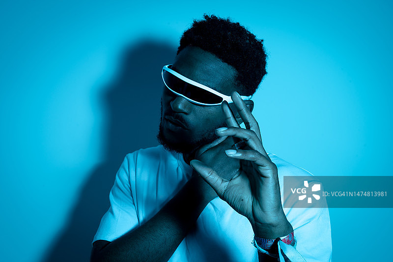 年轻的非裔美国男子戴着未来主义时尚眼镜，带有蓝色霓虹灯，用手势示意。未来概念、现代科技、技术进步、独眼巨人眼镜图片素材