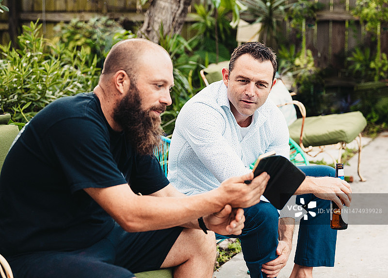男子向朋友展示手机，朋友们在户外泳池边休闲聚会，喝着饮料图片素材