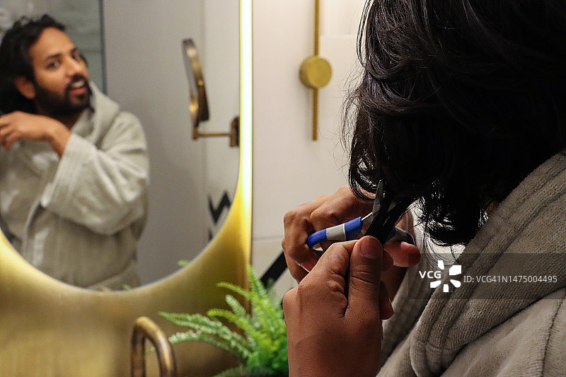 印度男人站在浴室水槽前，穿着浴袍，用剪刀修剪头发的镜子中的倒影特写，正在理发，金属盆中的人造蕨类植物，玻璃杯中的牙刷，自我护理和仪容的概念图片素材