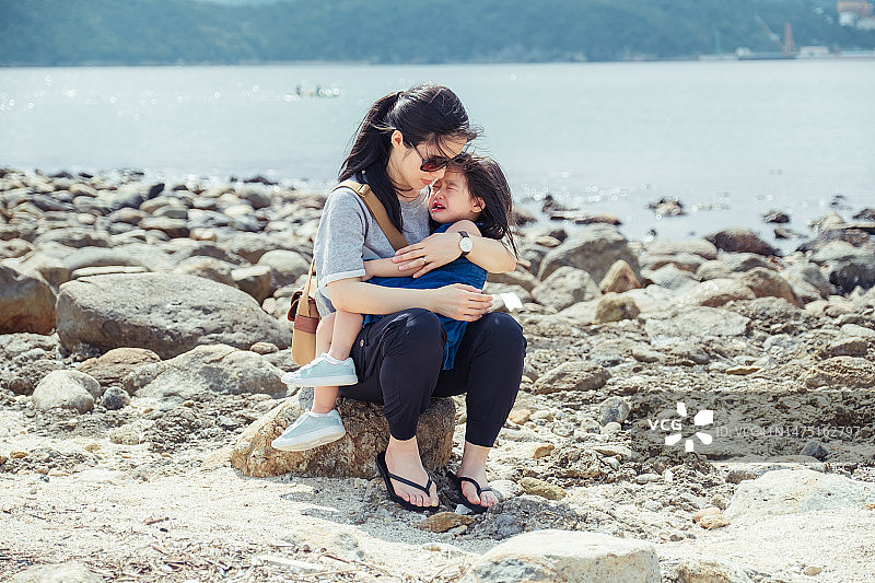 年轻的亚洲母亲抱着在海滩上受伤哭泣的小女儿，充满爱、关怀和母爱图片素材