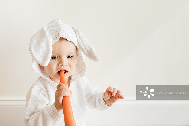 在自然光线下，一个15个月大的古巴裔美国男婴光着脚坐在地板上，第一次品尝胡萝卜图片素材