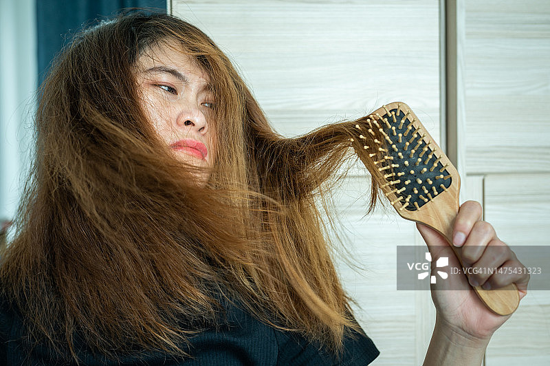 不快乐的亚洲女人在刷她凌乱和缠结的头发后感到压力。图片素材