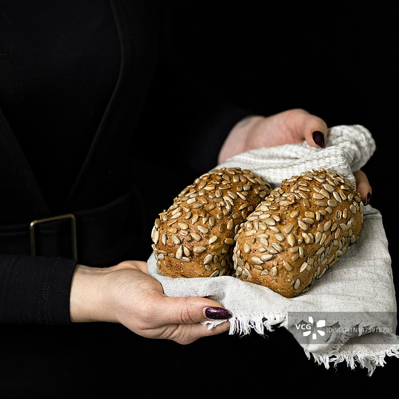 女人手中毛巾上的两块杂粮面包。含葵花籽面包。新鲜烘焙。健康饮食。黑色背景。侧视图。复制空间图片素材
