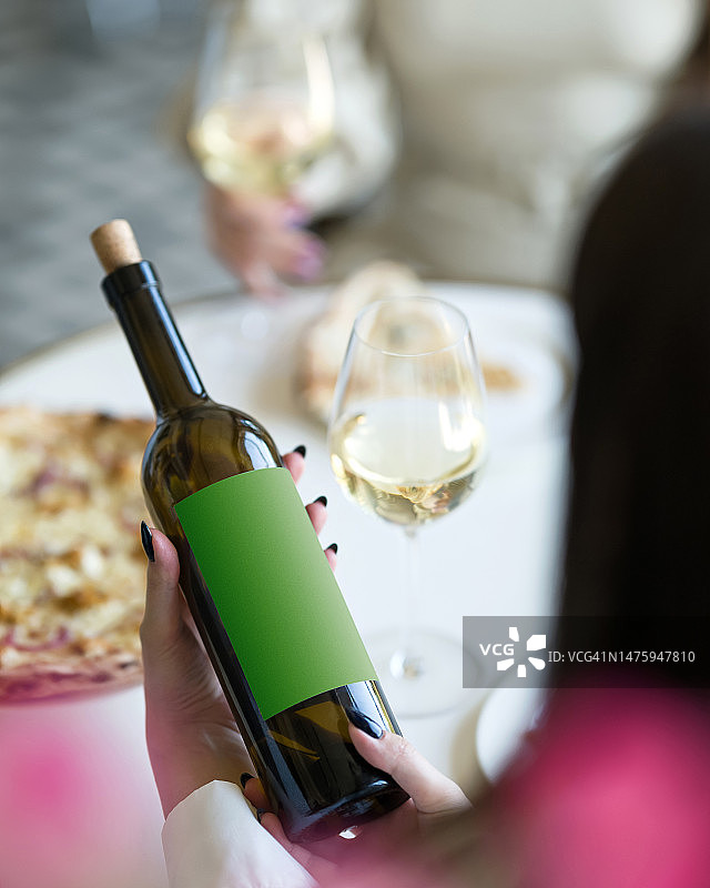 女人手握一瓶白葡萄酒。葡萄酒选择。阅读标签。玻璃杯中的白葡萄酒。两个女人喝酒吃披萨。品酒。模糊的背景。柔焦。侧视图图片素材