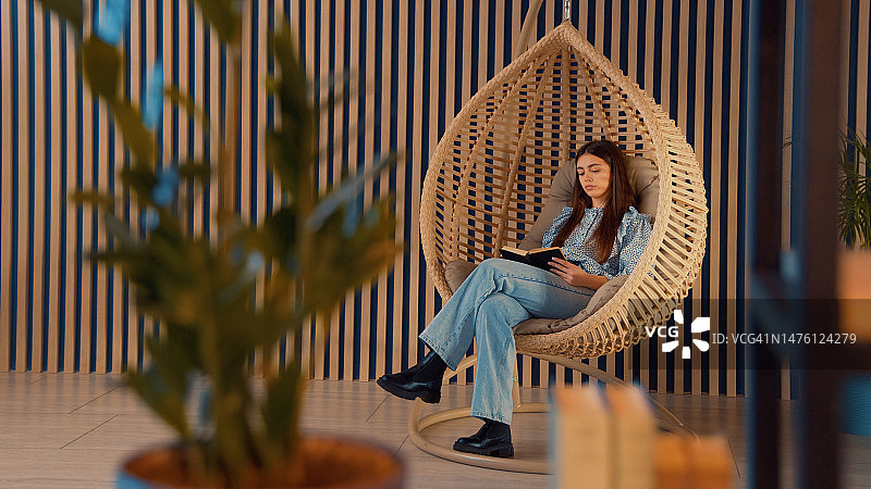 年轻女人坐在舒适的吊椅上读书，配有靠垫图片素材