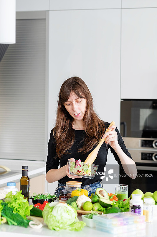 一位美丽苗条的女营养师穿着黑色T恤，在白色厨房的背景下，用木勺搅拌绿色沙拉图片素材