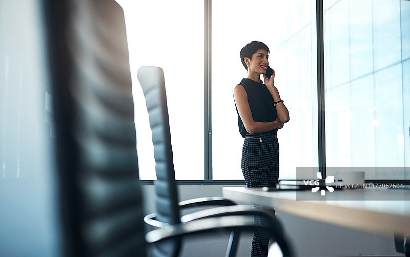 商务女性在会议室进行电话沟通，面带微笑讨论战略、公司事务或进行对话。快乐的女高管在办公室的窗边用智能手机洽谈公司业务图片素材