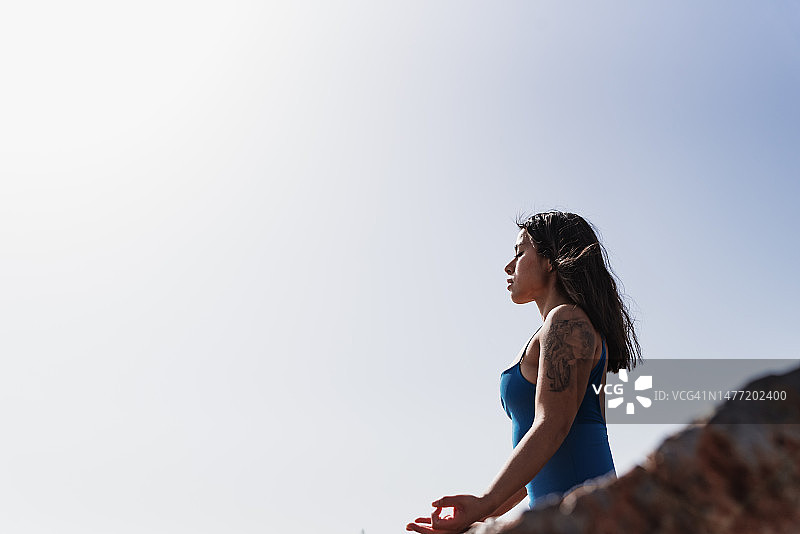 穿着蓝色紧身衣的年轻拉丁裔女性在海边的岩石上冥想图片素材